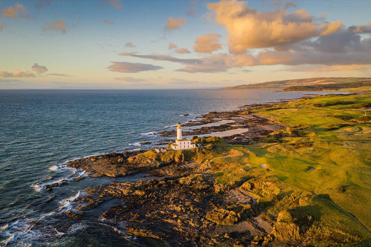 Aerial view of Turnberry lighthouse on its rocky point at golden hour, with the Ailsa course greens and bunkers on the right and the Ayrshire coast curving away behind